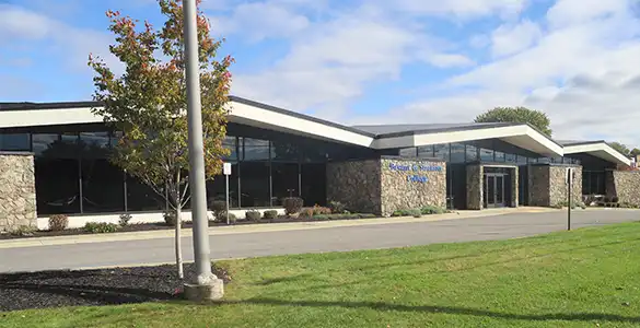 A modern, low-rise building with large windows and stone walls, surrounded by green grass and a tree in the foreground.