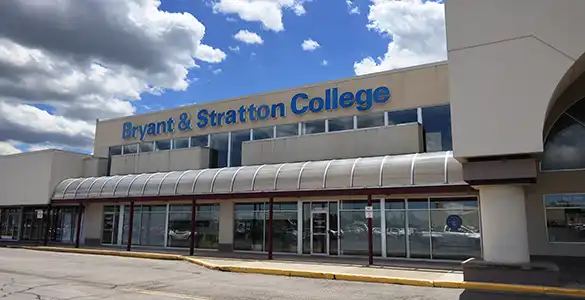 The exterior of Bryant & Stratton College building under a partly cloudy sky with a parking area in front.