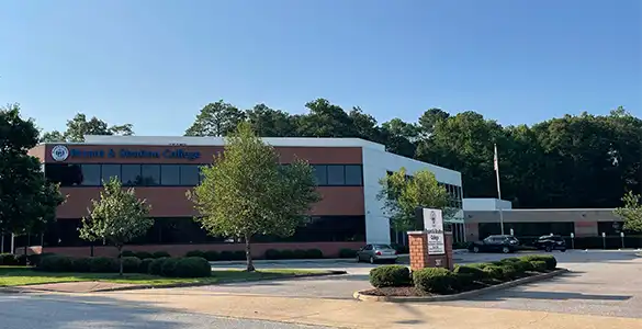 A two-story building with Bryant & Stratton College signage, surrounded by trees and a parking lot, on a sunny day.