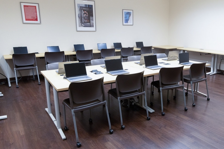 A classroom with wooden floors, several tables arranged in rows, brown chairs, and laptops on each table. Three framed pictures hang on the cream-colored walls.