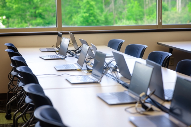 A row of open laptops on a long conference table in a meeting room with large windows overlooking trees and greenery outside in Orchard Park, NY.