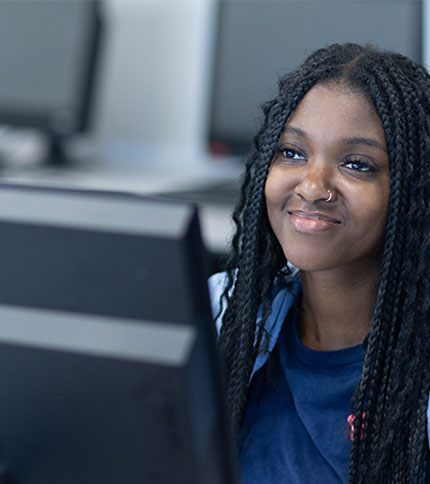 A person with long braided hair and a nose ring smiles while sitting in front of a computer.