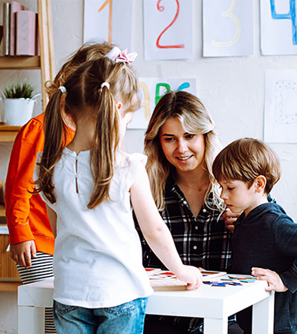 A woman sits at a table with three children engaged in an activity. They appear to be focused on something in front of them, with colorful papers and numbers on the wall in the background, suggesting a learning environment.