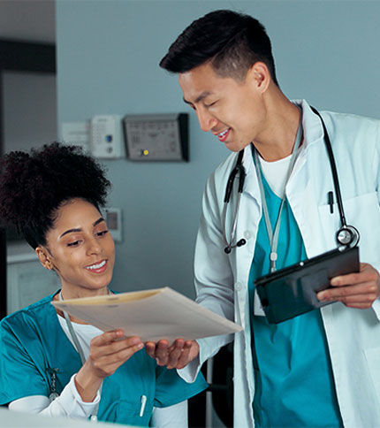 Two healthcare professionals in teal scrubs are reviewing documents together. A male doctor holds a tablet, while a female nurse looks at the papers. They are smiling and engaged in a well-lit medical office setting.