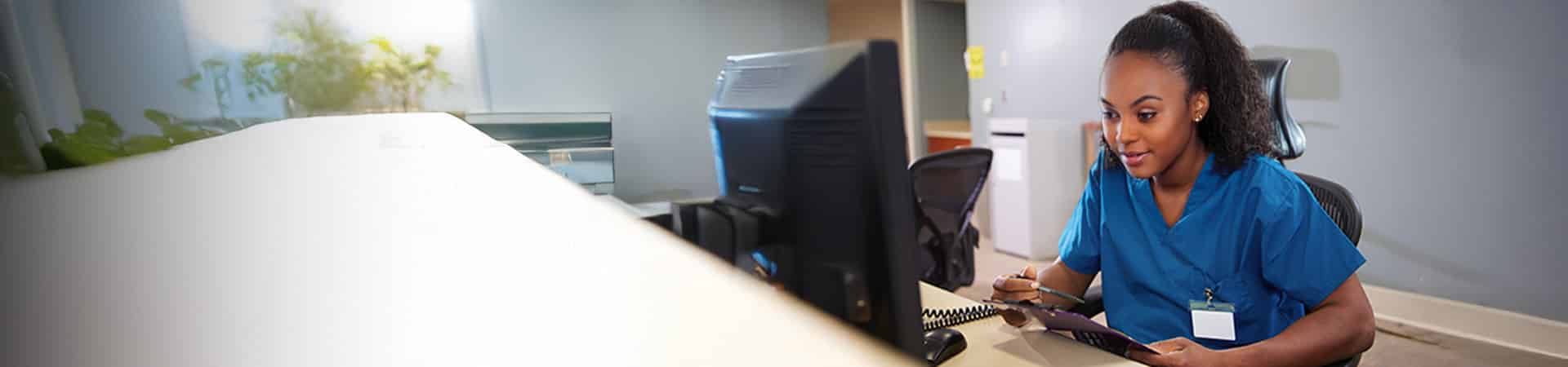 A woman in blue medical scrubs sits at a desk in a clinic, looking at a computer monitor and writing on a clipboard—an everyday scene for students enrolled in practical nursing programs in New York.