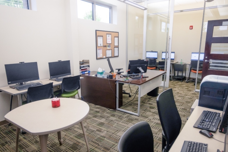 A small office in Orchard Park, NY, with several desks and computers, chairs, a round table, bulletin boards, and windows letting in natural light. The space is tidy and organized.