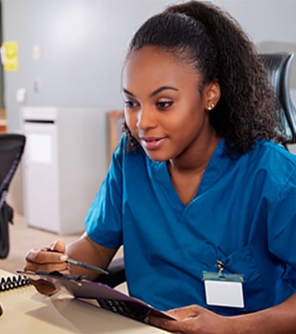 A woman in blue medical scrubs sits at a desk, looking at a tablet and holding a pen. With her name badge on, she appears focused—possibly studying for a nursing certificate or enrolled in practical nursing programs in New York.