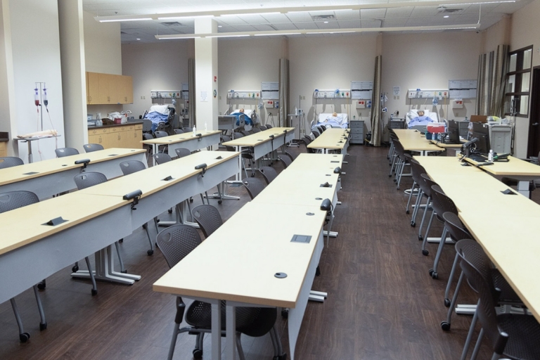 A classroom with rows of empty desks and chairs faces hospital beds along the walls, suggesting a medical training or simulation lab. The room is brightly lit with equipment and supplies visible near the beds.