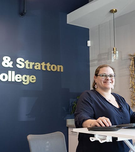 A person standing and smiling at a reception desk in front of a blue wall with "Stratton College" written on it. The space is modern, with a hanging light fixture and minimalistic decor.