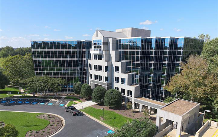 A modern glass office building with five stories, surrounded by trees and greenery, reflecting the blue sky—an ideal location for colleges in Richmond VA or nursing programs in Richmond, with a parking lot and a circular driveway at the entrance.