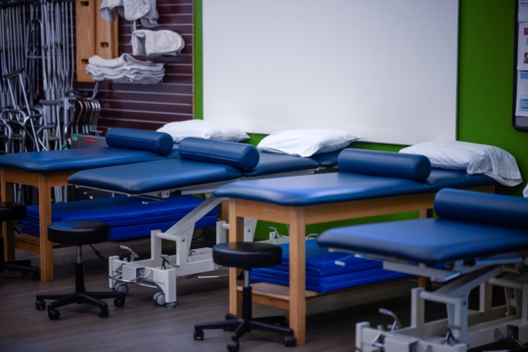 A row of blue padded physical therapy tables with pillows, stools, and folded towels in a medical clinic in Orchard Park NY, with mobility aids visible in the background.