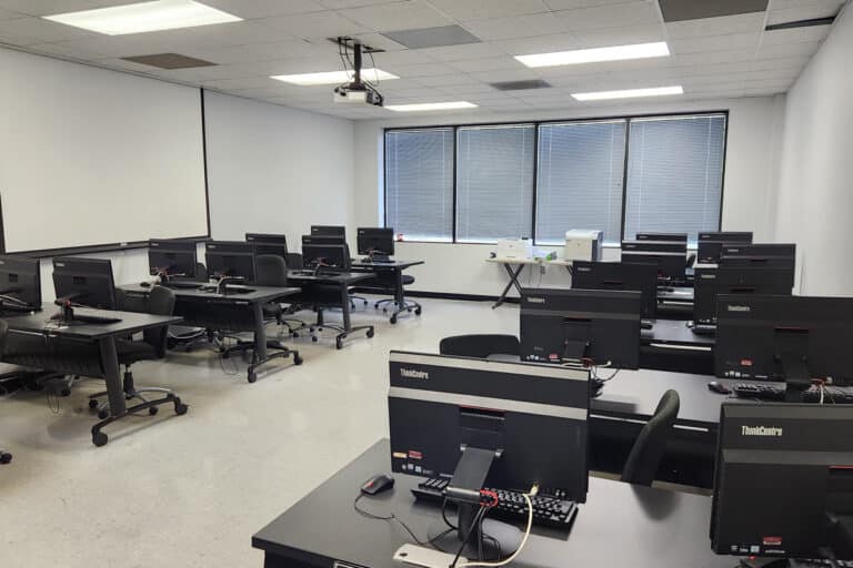 A computer lab with several desks, each equipped with a desktop computer, facing a large projection screen.