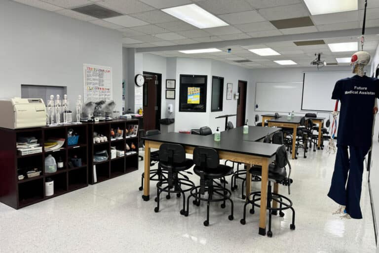 A medical assistant training lab with tables, chairs, anatomical models, and a mannequin wearing a 'Future Medical Assistant' shirt.
