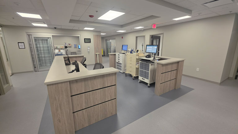 Nursing simulation lab with nurse station desks, computers, medical carts, supply cabinets, and clinical practice rooms.