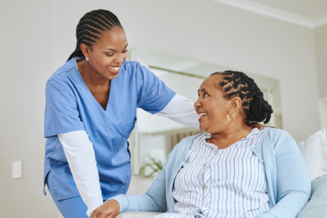 A nurse in blue scrubs, pursuing her RN to BSN, smiles and talks with an older woman sitting on a bed. The two women are making eye contact and holding hands in a bright, comfortable room.