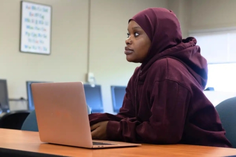 A woman wearing a maroon hijab and hoodie sits at a desk with a laptop, looking thoughtfully to the side in a classroom setting at one of the top colleges in Richmond, VA, with computers lining the background.