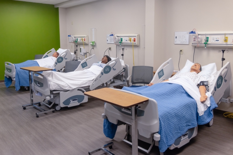 Three hospital beds with medical mannequins covered by blue blankets in a clinical training room in Orchard Park, NY, with medical equipment and supplies on the walls behind the beds.