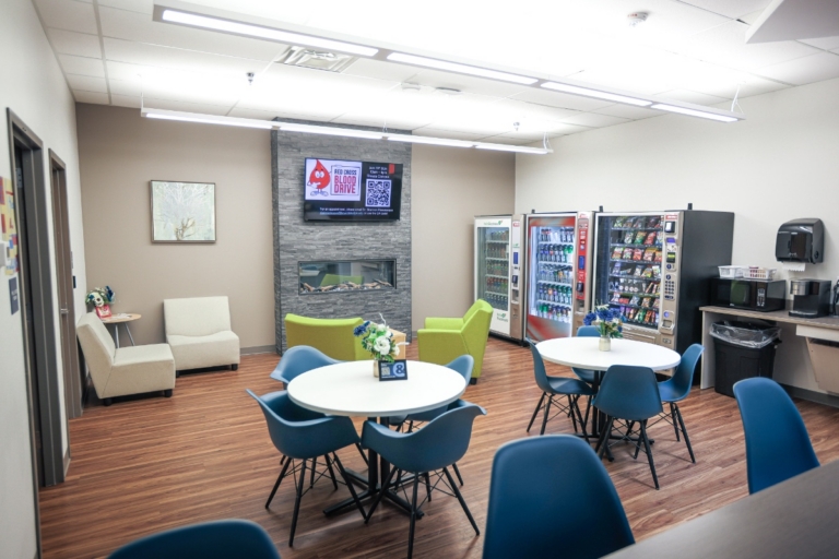 A break room in Henrietta, NY, with round tables, blue chairs, vending machines, a microwave, a coffee maker, armchairs, and a TV mounted above a fireplace. Wooden floor and bright lighting.