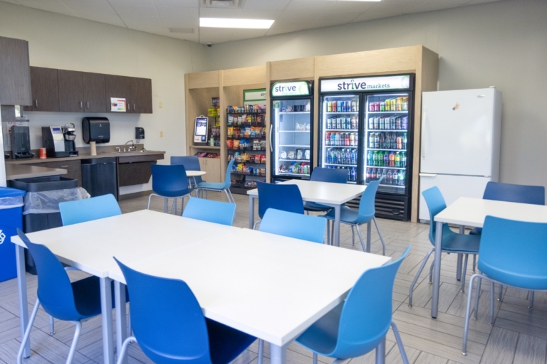 A break room in Orchard Park, NY, with white tables, blue chairs, vending machines stocked with snacks and drinks, a refrigerator, a sink, and a coffee maker. The room is well-lit and organized for employee use.