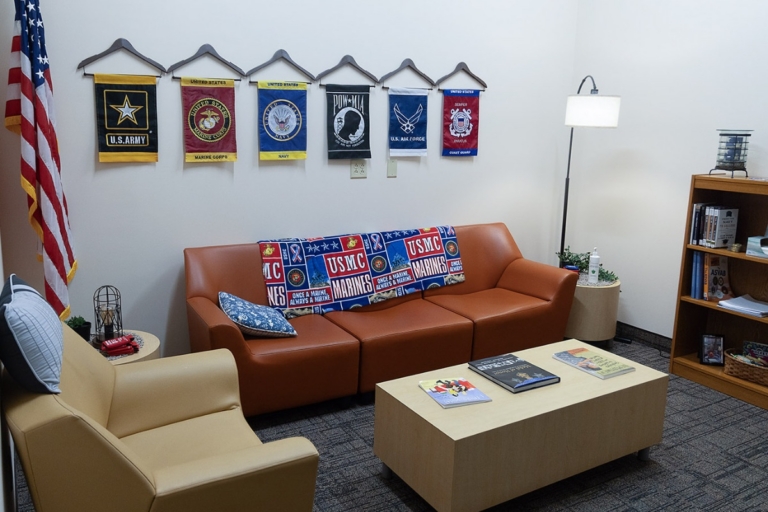 A cozy office with brown sofas, military flags on the wall, an American flag, bookshelves, a coffee table with magazines, and a floor lamp. The decor includes patriotic and military-themed items.