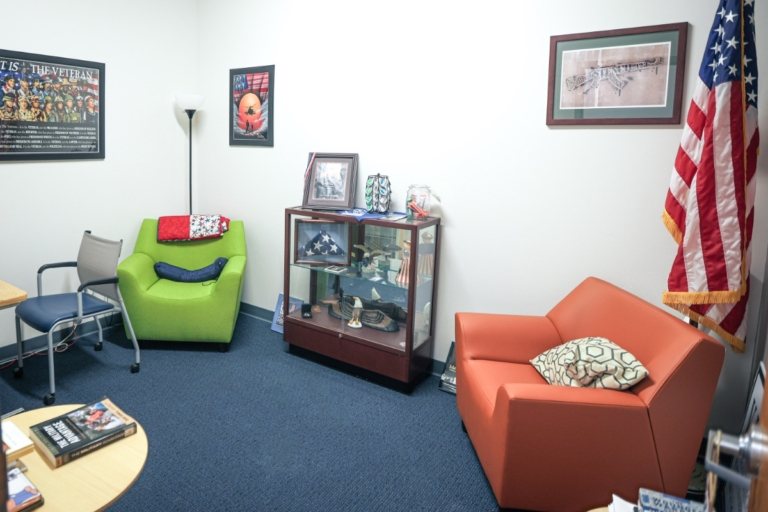 A small office in Henrietta, NY, with blue carpet, green and orange chairs, a display case with military memorabilia, framed art on white walls, and an American flag in the corner.