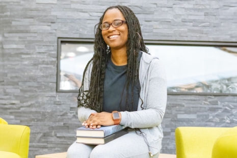 A woman with long braids, wearing glasses and a gray outfit, smiles while sitting indoors. She holds two books on her lap—perhaps researching nursing programs in Virginia Beach—and sits between two green chairs by a gray stone wall.