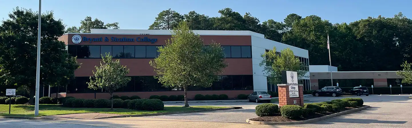 A brick and glass building of Bryant & Stratton College with a parking lot, trees, and a sign in front.