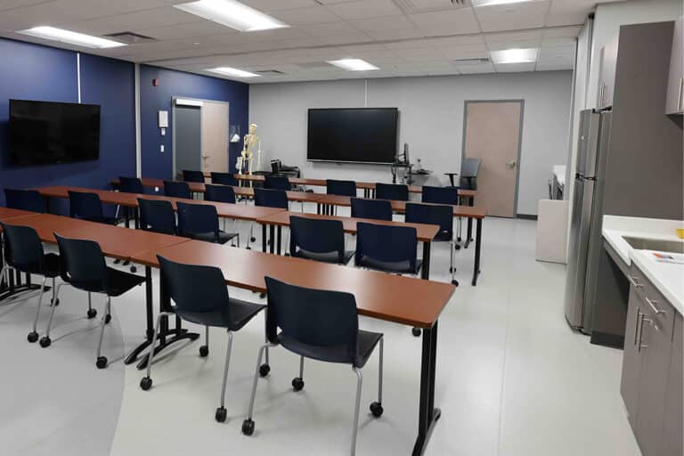 A modern classroom in Liverpool, NY, with rows of brown tables and blue chairs, a large TV screen and whiteboard at the front, a skeleton model by the wall, plus cabinets and a refrigerator on the right side.