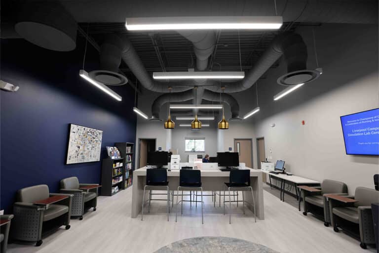 Modern office space in Liverpool, NY with blue and gray walls, desks with computers, chairs, bookshelves, a wall-mounted display screen, exposed ductwork, pendant lights, and a small window at the far end letting in natural light.