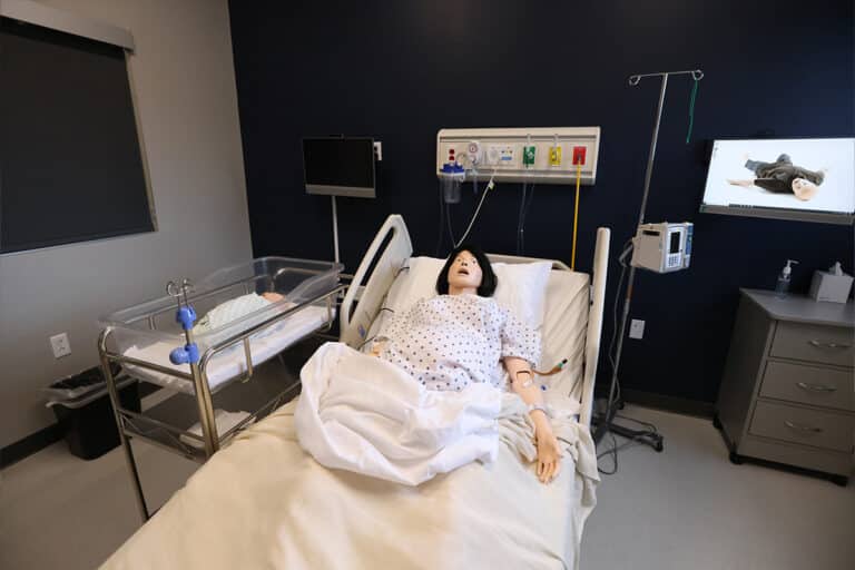 A medical simulation room in Liverpool, NY, features a life-like mannequin in a hospital bed, a baby crib beside it, medical equipment on the wall, and a monitor and IV stand nearby.
