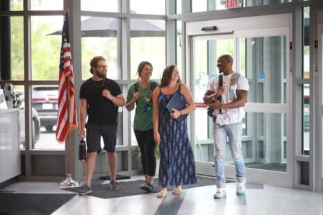 Four adults smile and chat as they walk into a modern building with large windows in Liverpool, NY. One carries a water bottle, another has a notebook, and there’s an American flag near the entrance.