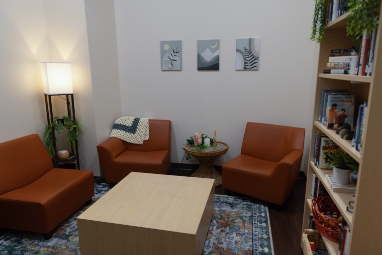 A cozy room with three orange chairs, a wooden coffee table, a floor lamp, bookshelves, plants, a round side table, and three minimalist art pieces on the wall.