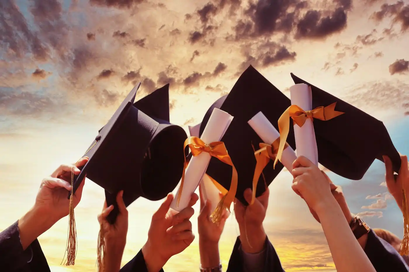 Graduates holding their caps and diplomas up in the air against a dramatic sunset sky.