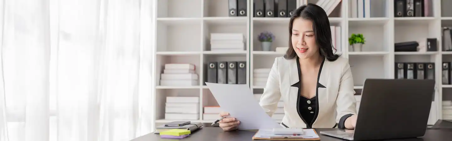 A woman in a business suit reviews a document at her desk with a laptop and binders in the background.