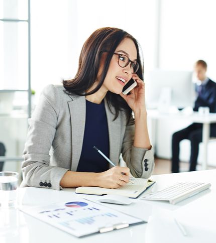 An Administrative Assistant in business attire sits at a desk, talking on a mobile phone, taking notes in a notebook, and smiling. A keyboard, documents, and a glass of water are on the desk, with another person in the background.