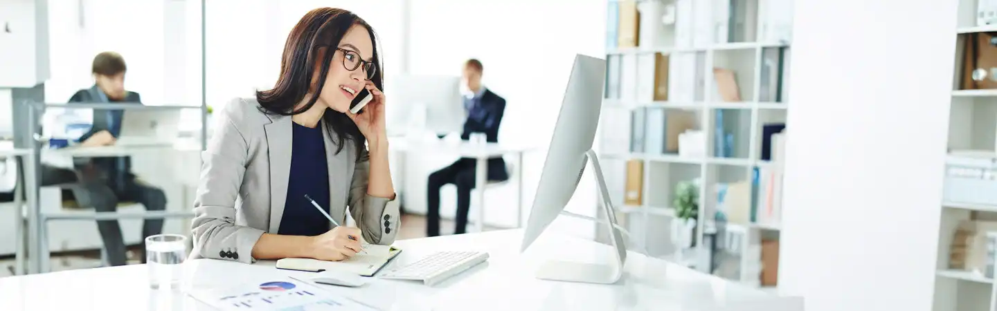 Woman in an office talking on the phone, writing on a notepad, and looking at a computer screen, with colleagues in background.