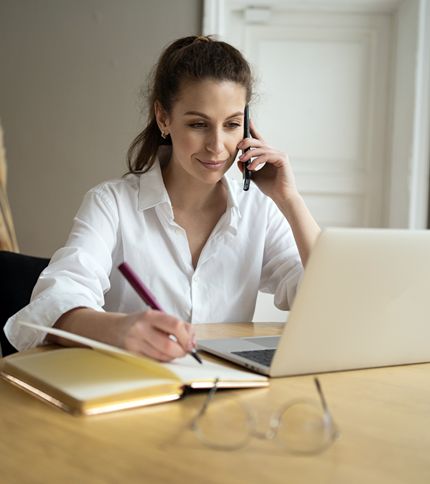 An Administrative Coordinator in a white shirt sits at a desk, talking on the phone, writing in a notebook, and working on a laptop. Glasses and a closed notebook rest on the table in front of her.