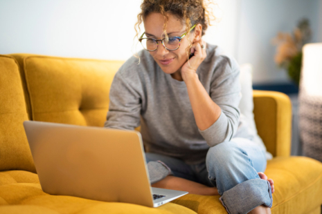 A woman with curly hair and glasses sits on a yellow sofa, smiling at her laptop as she researches nursing programs in Virginia Beach. Wearing a gray sweater and jeans, she appears relaxed and engaged with her work.
