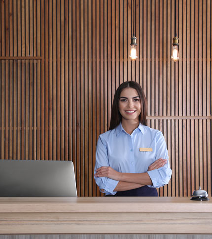 A smiling woman in a blue uniform stands with arms crossed behind a reception desk. A laptop and bell are on the desk. Wood panel wall and hanging light bulbs are in the background.