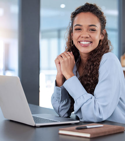A person with long curly hair smiles while sitting at a desk with a laptop and a notebook. They are wearing a light blue shirt and appear to be in a modern office setting.