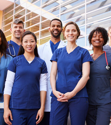 A group of healthcare professionals, wearing blue and white medical scrubs, stand smiling in front of a modern building with large windows.
