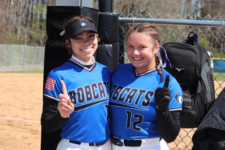 Two softball players in blue "Bobcats" jerseys, smiling and holding up one finger each near a chain-link fence on a sunny day.