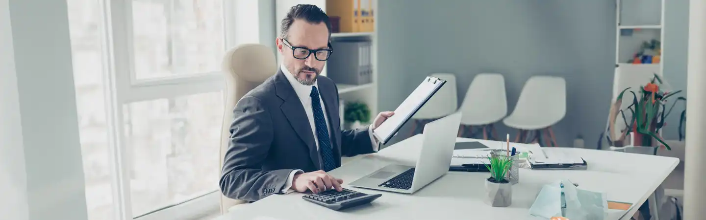 A man in a suit works at a desk with a laptop, holding a clipboard and using a calculator in a modern office.
