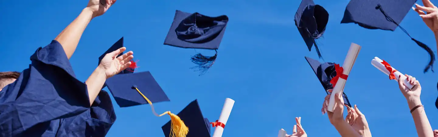 Graduation caps and diplomas are joyfully tossed in the air against a clear blue sky in celebration.