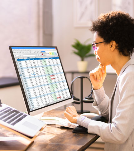 A person wearing glasses is working at a desk, looking at a computer monitor displaying a spreadsheet. A laptop and plant are also on the desk. The setting appears to be a bright, modern office.