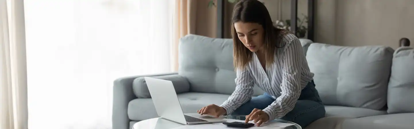 A woman sits on a couch using a laptop while reaching for a calculator on a table in front of her.