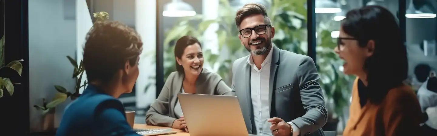 A group of four people in business attire, sitting in an office, smiling and having a discussion with a laptop on the table.