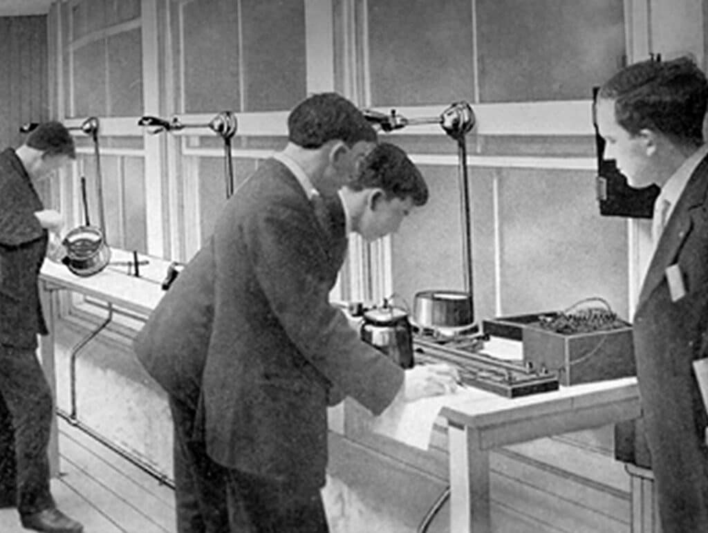Four men conducting experiments at lab tables near large windows, adjusting scientific equipment and taking notes.