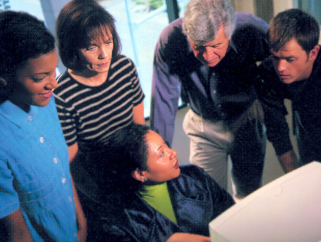 Five people gathered around a computer, looking at the screen together in a professional setting.