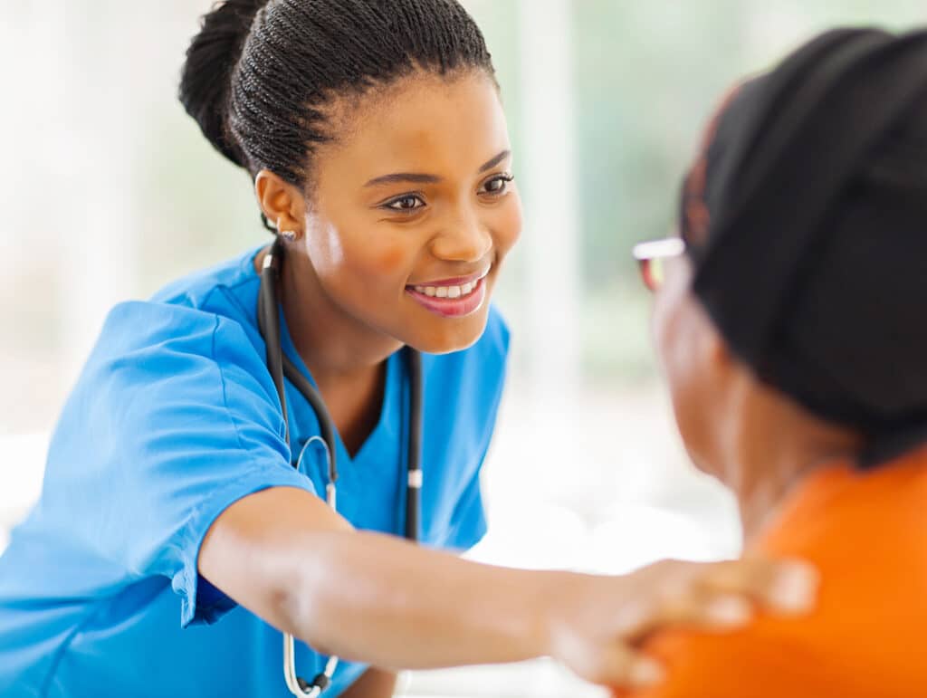 A nurse in blue scrubs with a stethoscope comforts an elderly patient by gently holding their shoulder.
